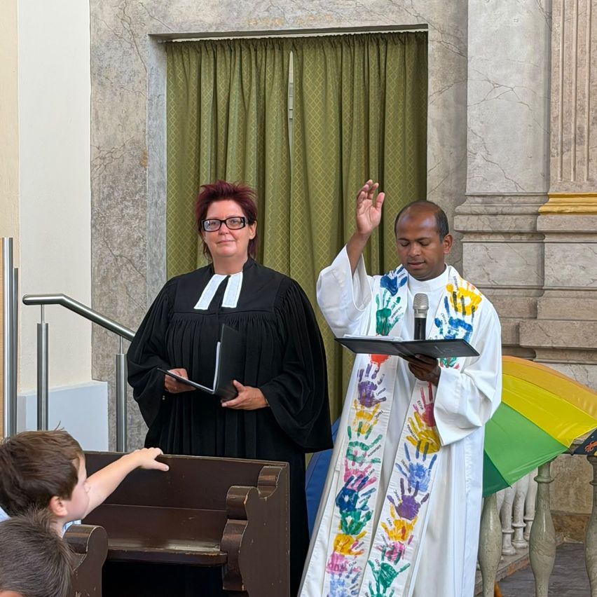 Ein Priester und eine Frau in einem schwarzen Gewand stehen vor einem Gebäude. Der Priester hält ein Buch und ein Mikrofon. Ein Junge sitzt auf einer Bank.