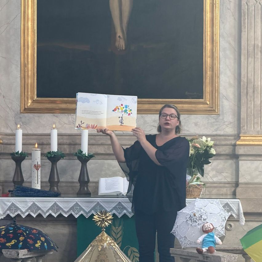 Eine Frau steht in einer Kirche und hält ein Buch in der Hand, das sie laut liest. Hinter ihr hängt ein großes Gemälde, und vor ihr steht ein Tisch mit Kerzen und Blumen.