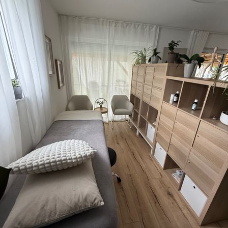 Ein Innenbereich mit einem Massagetisch, Kissen und Stühlen. Es hat Holzböden und weiße Vorhänge. Es gibt einen Schrank mit Regalen und Schubladen, der verschiedene Gegenstände und Topfpflanzen beherbergt.