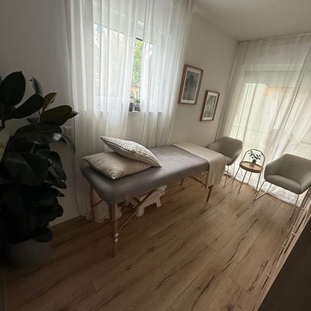 Ein Raum mit Holzboden, einem Massagetisch, zwei grauen Stühlen und einem Topfpflanzen. Die Wände sind weiß mit Vorhängen, und es gibt zwei gerahmte Bilder an der Wand. Ein Fenster ist hinter den Vorhängen sichtbar.