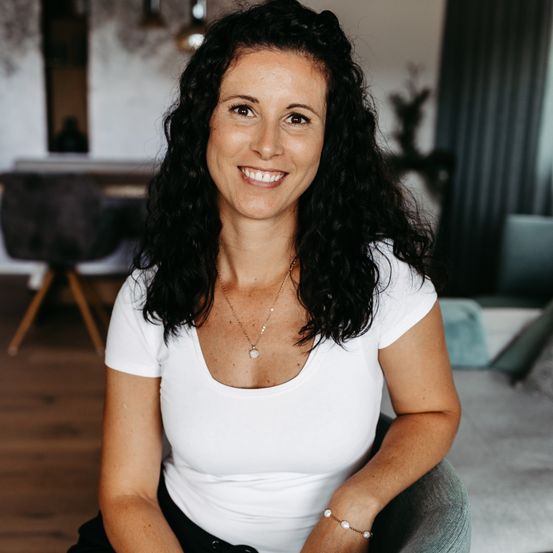 Eine Frau mit lockigem schwarzem Haar lächelt und sitzt auf einem Stuhl. Sie trägt ein weißes T-Shirt, eine Halskette und ein Armband. Hinter ihr ist eine verschwommene Ansicht eines Tisches und Stühle zu sehen.