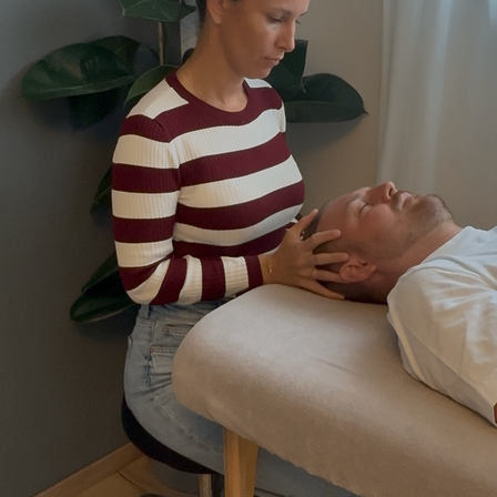 Eine Frau massiert den Kopf eines Mannes mit ihren Händen. Der Mann liegt auf einem weißen Bett. Die Frau trägt ein gestreiftes Shirt und Jeans. Hinter ihr steht eine Pflanze in einem Topf.