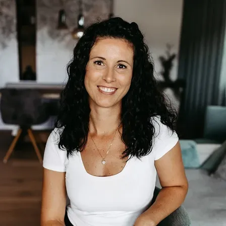 Eine lächelnde Frau mit lockigem Haar, die ein weißes Oberteil und schwarze Hose trägt, sitzt auf einem Stuhl in einem Raum mit Holzboden.