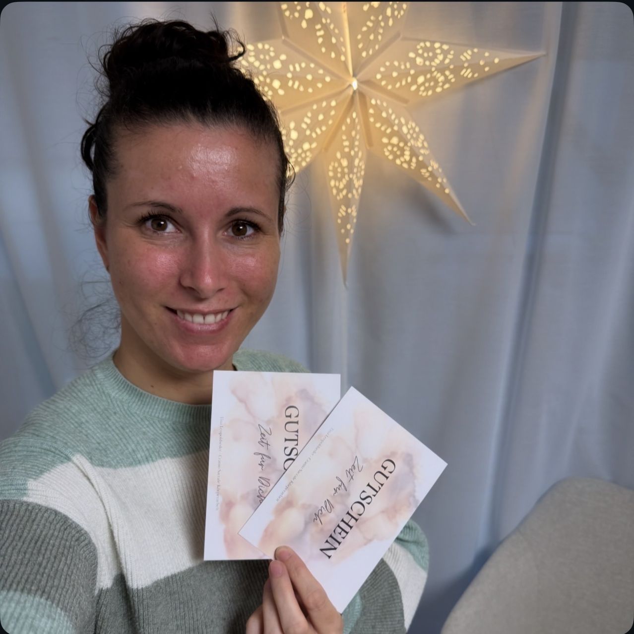A woman holds two cards with 'Gutschein' written on them. She is smiling and posing against a white backdrop with a star-shaped light in the background.