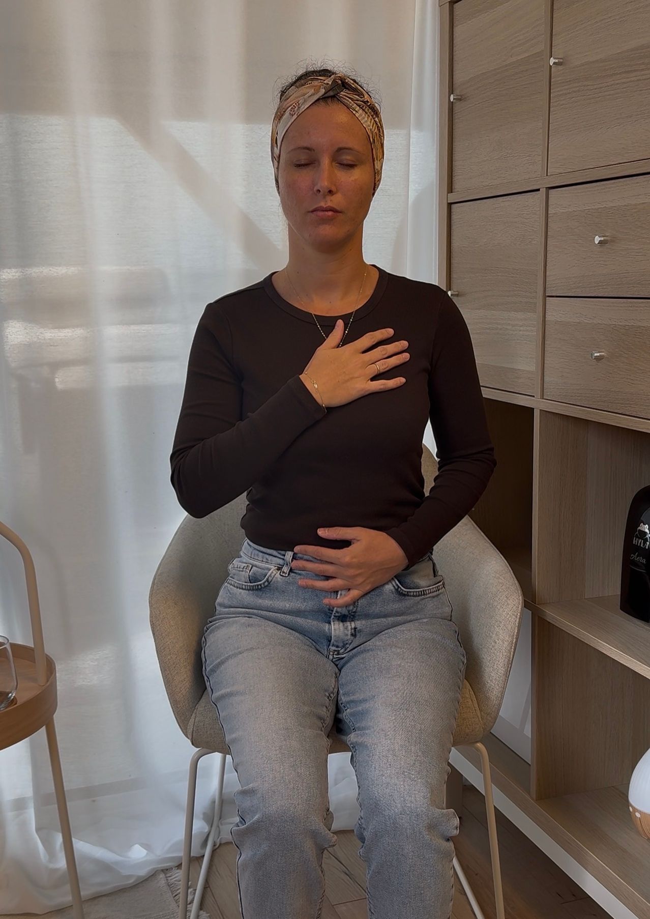 Eine Frau sitzt auf einem Stuhl, trägt ein schwarzes Shirt und Jeans. Ihre Hände liegen auf Brust und Bauch, und ihre Augen sind geschlossen. Im Hintergrund befindet sich ein Schrank mit Schubladen.