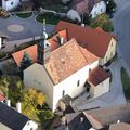 Luftaufnahme einer Kirche mit goldenem Turm und roten Dächern, eingebettet zwischen anderen Häusern mit Bäumen und einer asphaltierten Straße.