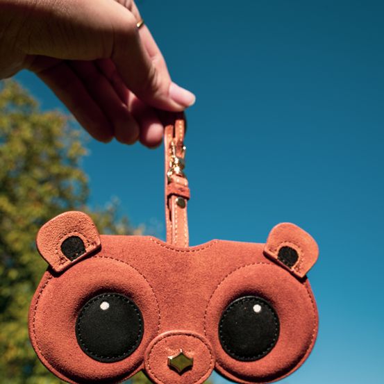 Eine Person hält eine braune, bärenförmige Geldbörse im Freien. Die Geldbörse hat einen goldenen, sternförmigen Anhänger und große schwarze Augen. Der Himmel ist klar und blau.