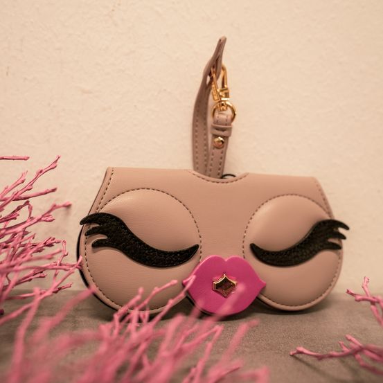 Eine beige Handtasche mit rosa Lippen und schwarzen Wimpern liegt auf einer grauen Oberfläche mit rosa Zweigen.