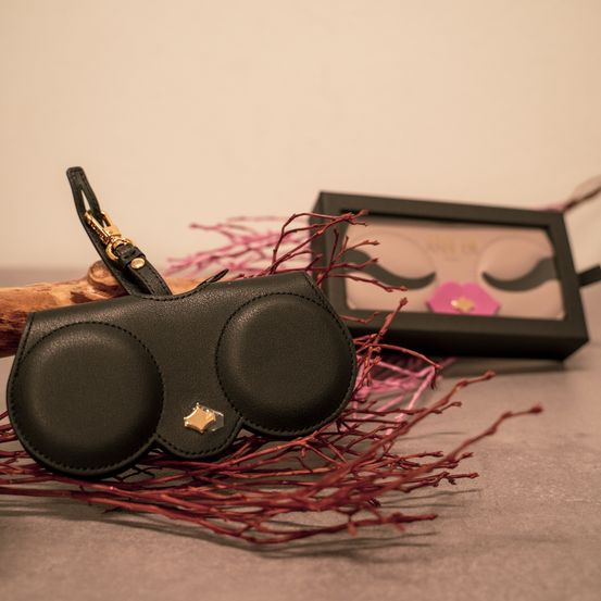 Ein schwarzer lederähnlicher Gegenstand, der an eine Brille erinnert und goldene Akzente hat, liegt auf roten Zweigen, dahinter ein schwarzer Bilderrahmen.