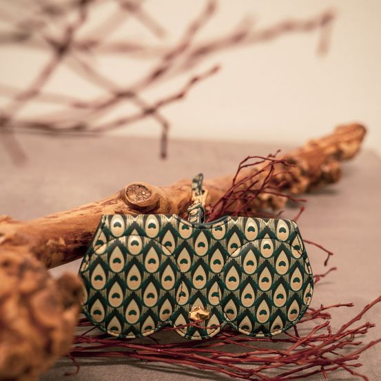 Eine grün-goldene Clutch-Tasche ruht auf einem Ast mit roten Zweigen auf einer grauen Oberfläche.