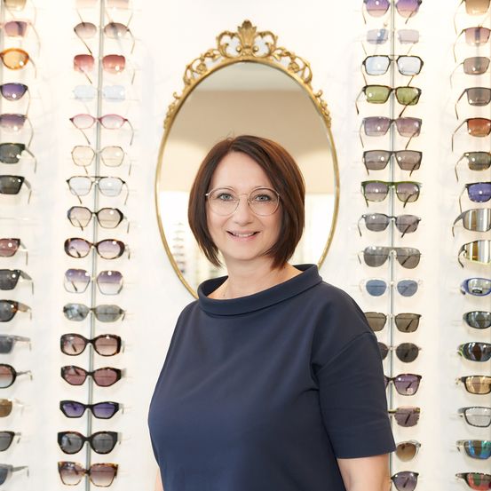 Eine Frau in einem marineblauen Oberteil steht vor einer Wand mit verschiedenen Sonnenbrillen. Sie lächelt und trägt eine Brille.