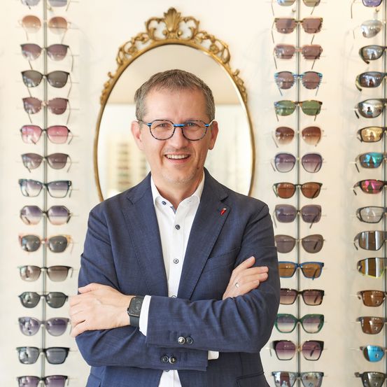 Ein Mann in einem blauen Anzug und mit Brille steht mit verschränkten Armen vor einer Wand mit Sonnenbrillen. Hinter ihm befindet sich ein Spiegel mit goldenem Rahmen.