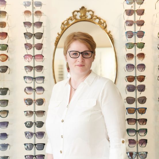 Eine Frau mit Brille steht vor einer Wand mit verschiedenen Sonnenbrillen. Sie lächelt und schaut direkt in die Kamera.