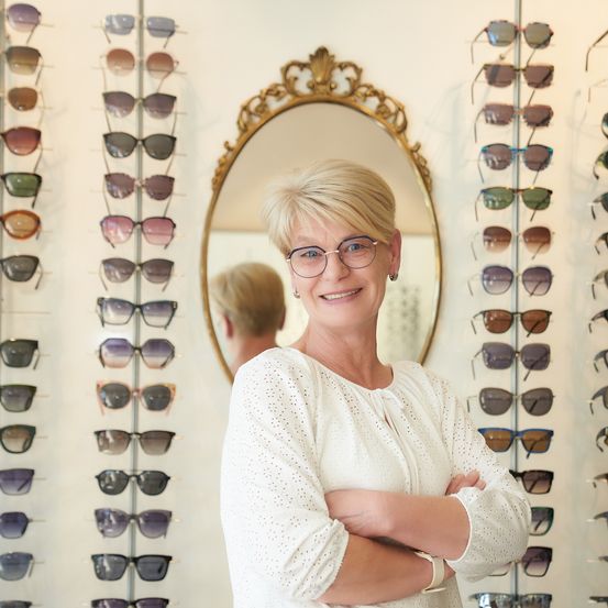 Eine Frau mit Brille steht selbstbewusst vor einer Wand mit verschiedenen Sonnenbrillen und reflektiert ihr Lächeln in einem Spiegel.