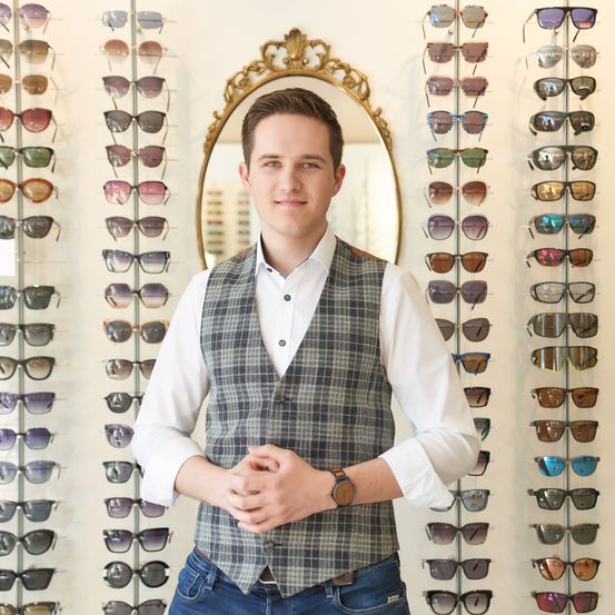 Ein Mann steht vor einer Wand voller Sonnenbrillen. Er trägt eine karierte Weste über einem weißen Hemd, mit einer Uhr an seinem linken Handgelenk.