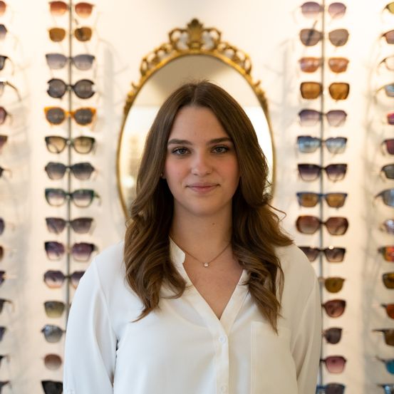 Eine Frau in einem weißen Hemd steht vor einem Spiegel und lächelt, mit vielen Paaren von Sonnenbrillen, die an der Wand hinter ihr ausgestellt sind.