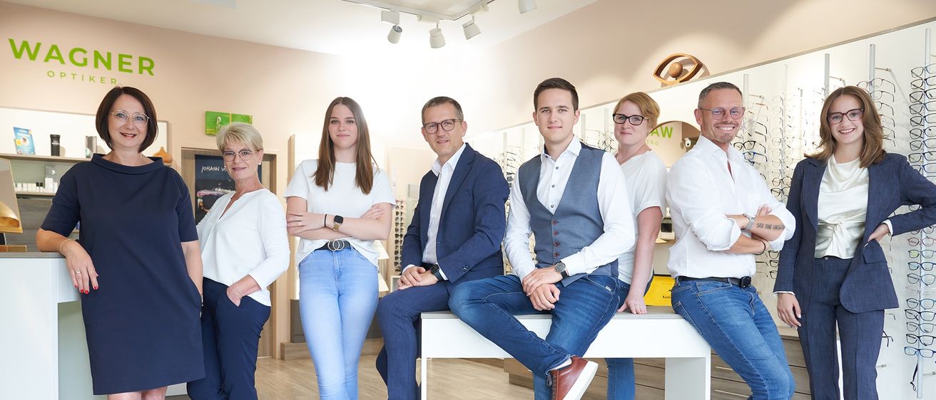 A group of people in professional attire are posing for a photo in a well-lit office. Some are seated on a table, while others stand behind. The setting includes a wooden floor and decorative elements.