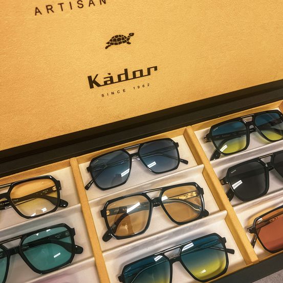 A box of sunglasses with the brand name 'Kador', established in 1962, is displayed. Various pairs of sunglasses are shown in the compartments of the box.
