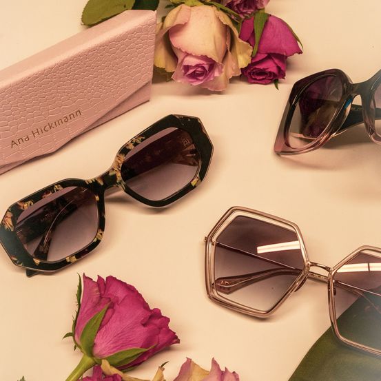 A set of sunglasses with floral designs and a pink box labeled Ana Hickmann are placed on a white surface with pink roses and green leaves.