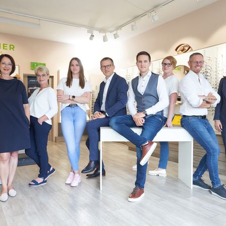 Eine Gruppe von Menschen posiert für ein Foto in einem Geschäft. Vier Männer sitzen auf einer Bank und drei Frauen stehen hinter ihnen. Sie tragen alle eine Brille und lächeln.