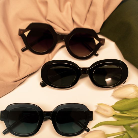 Three pairs of black sunglasses with slightly curved lenses are displayed on a white surface, surrounded by white tulips.