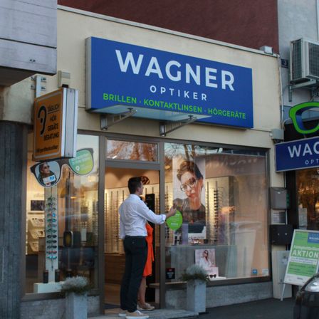Ein Optikerladen mit einem blauen Schild, das Wagner Optiker steht. Ein Mann und eine Frau stehen am Eingang.