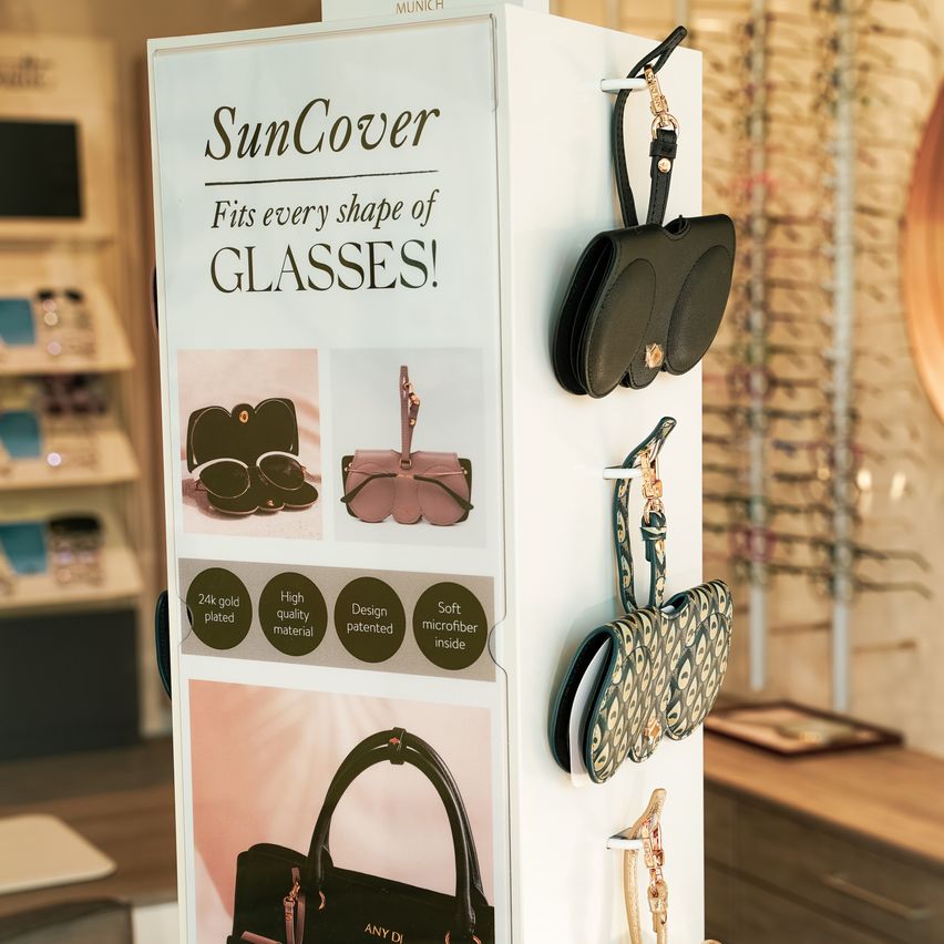 A white display board shows SunCover glasses cases with various designs and features such as 24k gold plating and soft microfiber inside.