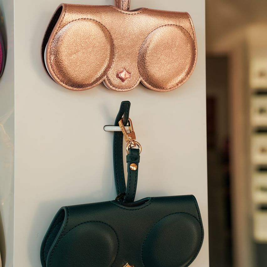 Two leather cases, one gold and one black, hang on a white wall. The gold case has a rose-gold charm.