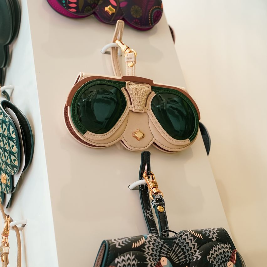 A collection of designer sunglasses and handbags displayed on a white wall. The sunglasses have green lenses and gold accents. The handbags feature various colorful designs and patterns.