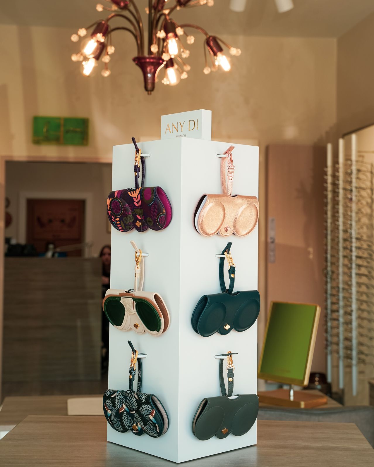 A display in a boutique showcases various designer glasses cases in diverse colors, hanging from a white stand with a sign that reads 'ANY DI'. Behind it is a room with a chandelier, a monitor, and a person in the distance.