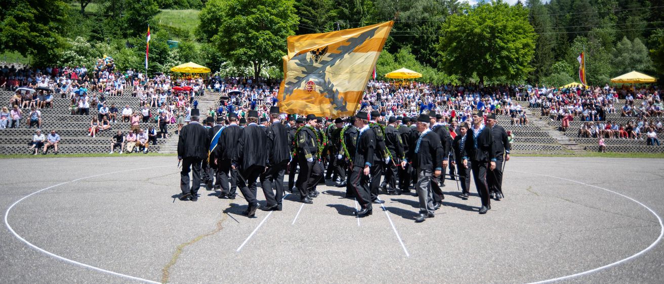 Eine zeremonielle Parade findet im Freien statt. Viele Menschen tragen schwarze Uniformen und halten eine Fahne. Sie marschieren im Kreis. Viele Menschen schauen zu. Bäume sind im Hintergrund.