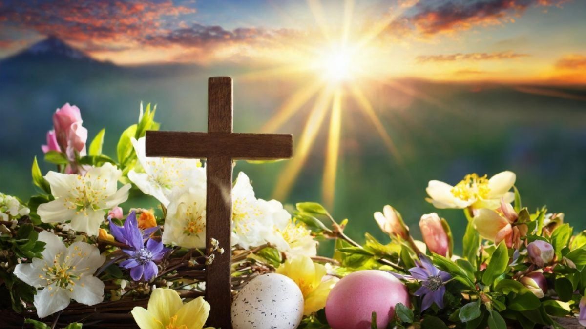 Ein hölzerne Kreuz wird von bunten Blumen und Ostereiern im Sonnenlicht umgeben.