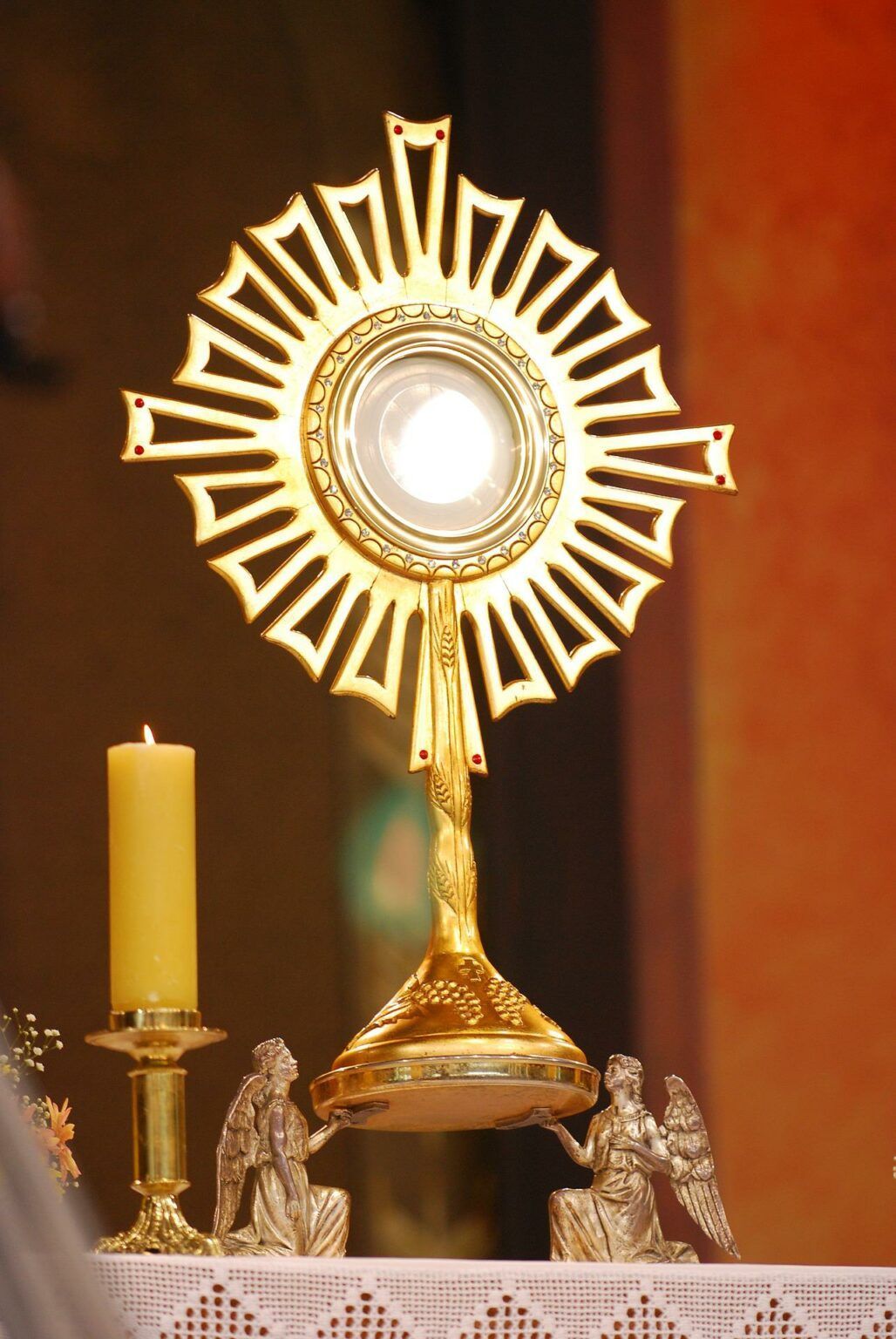Ein goldener Monstranz mit einer strahlenden Lichtquelle, begleitet von einer brennenden Kerze, steht auf einer goldenen Basis mit Cherubinenfiguren.