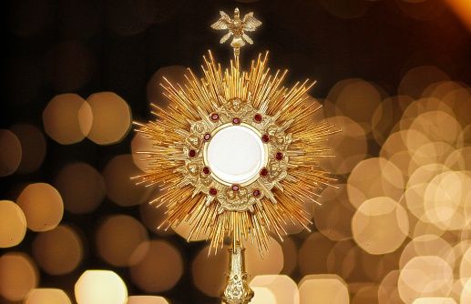A golden monstrance with a white circular object in the middle, surrounded by red jewels, is displayed against a blurred background of yellow lights.
