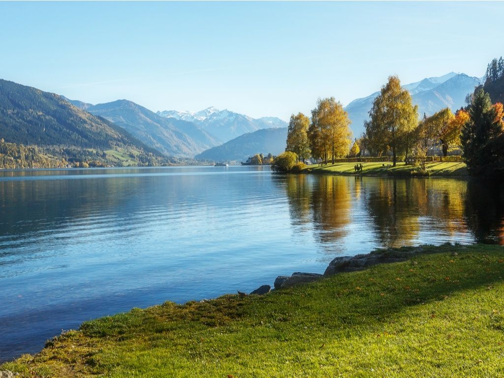 Herbstliche Bäume spiegeln sich im Zeller See, dahinter Berge
