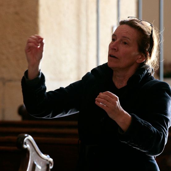 Eine ältere Frau steht in einer Kirche, trägt eine schwarze Jacke und hat eine Brille auf dem Kopf. Sie gestikuliert mit den Händen, möglicherweise dirigiert sie.