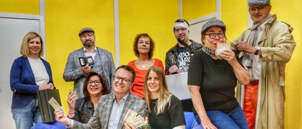 Eine Gruppe von Menschen posiert für ein Foto in einem Raum mit gelben Wänden. Eine Frau in der Mitte hält ein Getränk und etwas Geld, während ein Mann links von ihr einen Aktenkoffer hält.