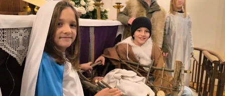 Vier Kinder in Kostümen stellen die Weihnachtskrippe in einer Kirche nach, wobei ein Mädchen in einem blauen Kleid Maria spielt, ein Junge mit Hut Josef und zwei andere Kinder als Hirten.