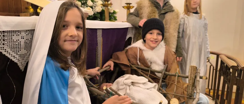 Vier Kinder in Kostümen stellen die Weihnachtskrippe in einer Kirche nach, wobei ein Mädchen in einem blauen Kleid Maria spielt, ein Junge mit Hut Josef und zwei andere Kinder als Hirten.