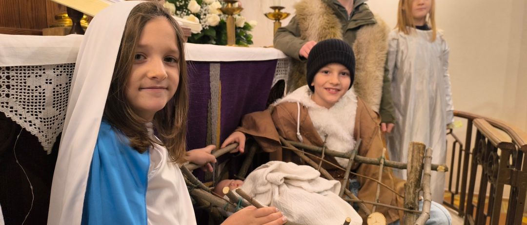 Vier Kinder in Kostümen stellen die Weihnachtskrippe in einer Kirche nach, wobei ein Mädchen in einem blauen Kleid Maria spielt, ein Junge mit Hut Josef und zwei andere Kinder als Hirten.