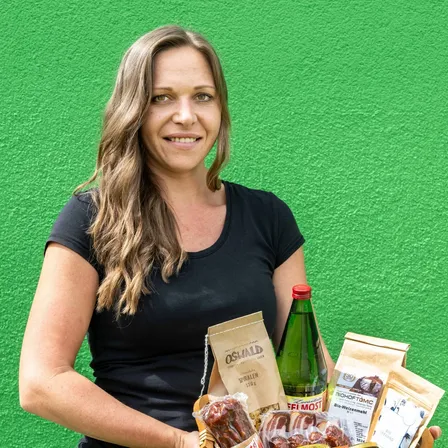 Eine Frau mit langen blonden Haaren, die ein schwarzes T-Shirt und blaue Jeans trägt, hält einen Korb voller verschiedener Lebensmittel, darunter eine Flasche Wein, eine Tüte Chips und ein Glas Marmelade.