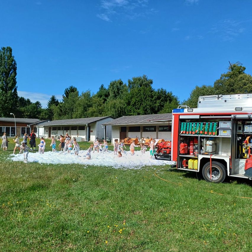 Eine Gruppe von Kindern spielt im Schaum, der von einem Feuerwehrauto auf einer Wiese mit Gebäuden und Bäumen im Hintergrund erzeugt wurde.
