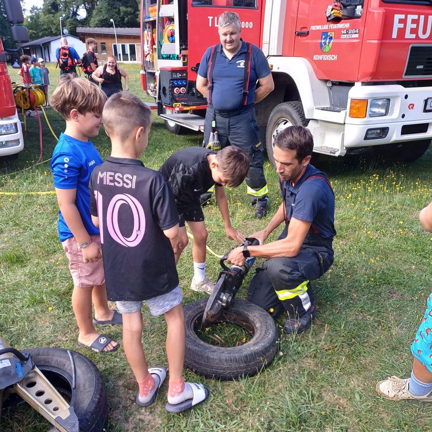Ein Feuerwehrmann demonstriert Kindern die Verwendung eines Reifenschneiders. Die Szene spielt sich draußen ab, mit einem Feuerwehrauto im Hintergrund.