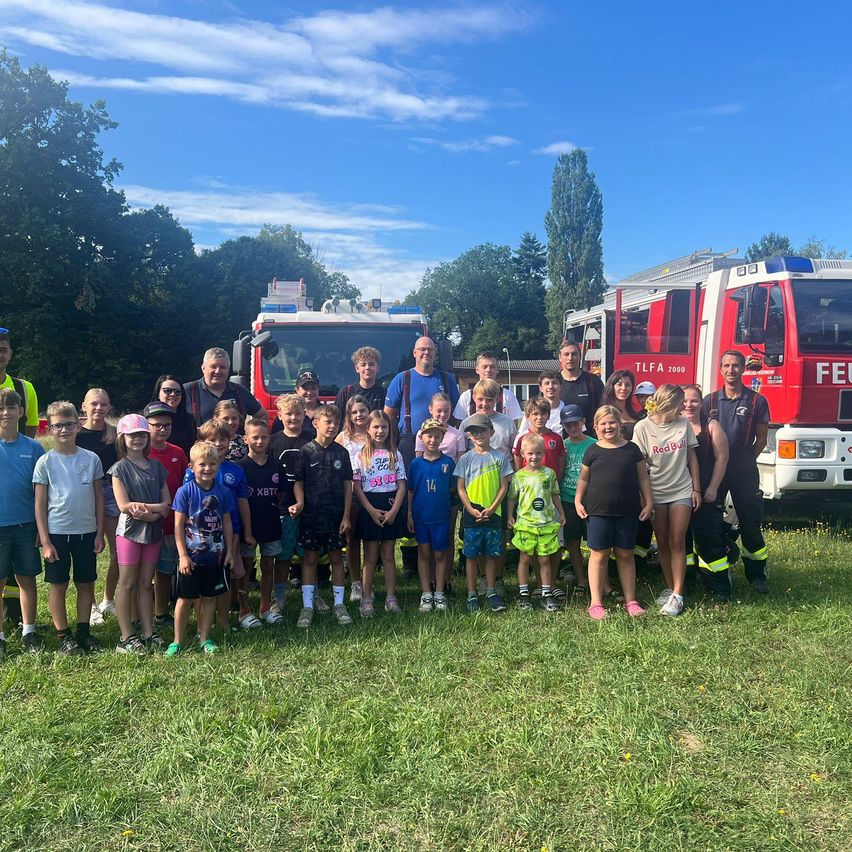 Eine Gruppe von Kindern und Erwachsenen posiert für ein Foto vor zwei Feuerwehrautos an einem sonnigen Tag.