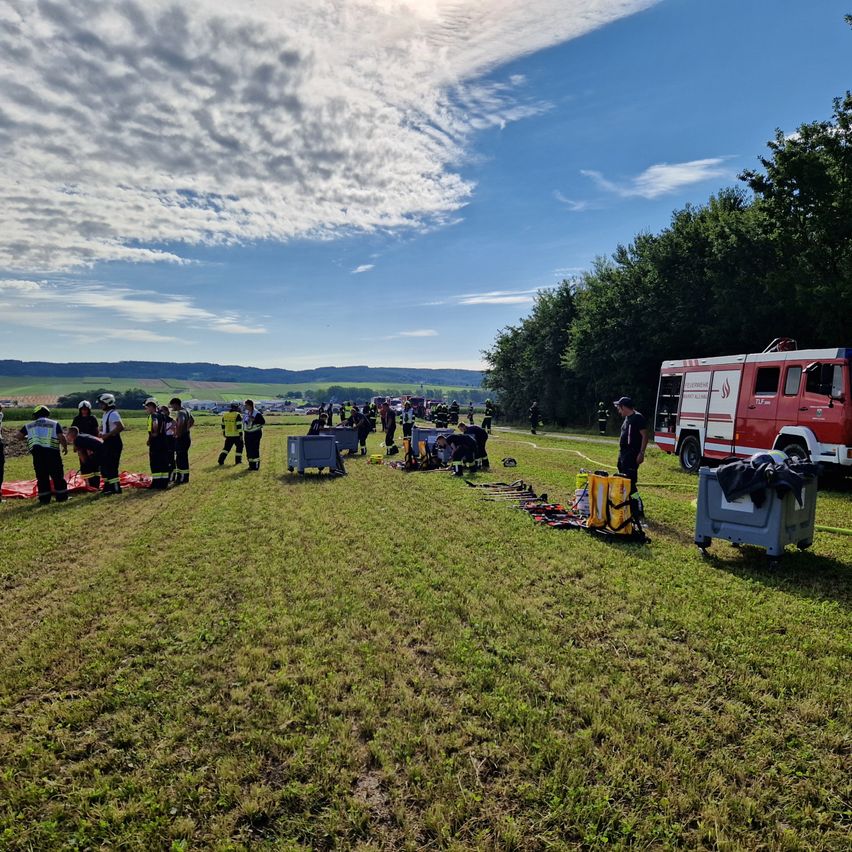Eine Gruppe von Menschen in Schutzkleidung versammelt sich auf einem Grasfeld, einige stehen, andere kümmern sich um Personen am Boden. In der Nähe befindet sich ein Feuerwehrauto und eine Person in der Nähe eines Trolleys. Die Szenerie ist vor einem Hintergrund von Bäumen und einem blauen Himmel mit Wolken.