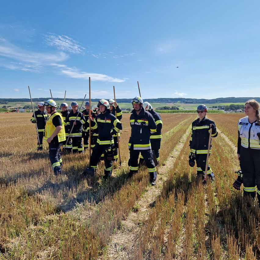 Eine Gruppe von Feuerwehrleuten in voller Ausrüstung, einschließlich Helmen und Stöcken, steht auf einem trockenen Feld unter einem blauen Himmel mit Wolken. Sie bereiten sich wahrscheinlich auf eine Übung oder Ausbildung vor.