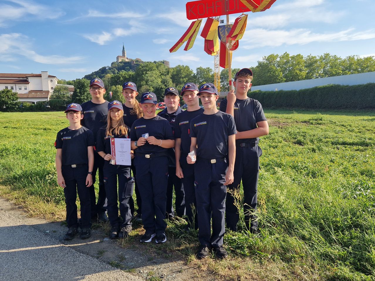 Eine Gruppe junger Menschen in dunkelblauen Uniformen steht auf einem Feld mit einem Schild hinter sich, das 'KOHFFELD' steht, und posiert für ein Foto.