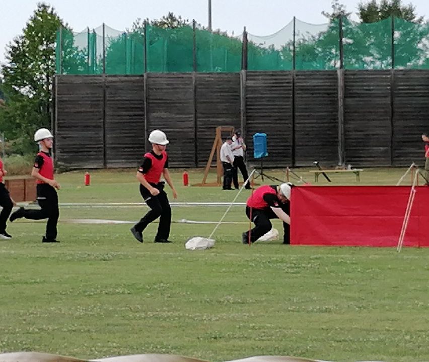 Eine Gruppe von Menschen in roten Westen und weißen Helmen läuft auf einem Feld mit einer roten Barriere davor. Eine Person hockt sich hinter die Barriere.