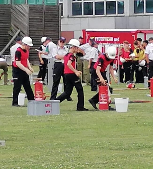 Mehrere Personen in roten und schwarzen Uniformen und weißen Helmen versammeln sich auf einem Feld, möglicherweise zur Übung eines Löscheinsatzes. Feuerlöscher stehen auf dem Boden. Dahinter stehen andere und beobachten. Ein rotes Fahrzeug mit dem Wort "Feuerwehr" ist geparkt.