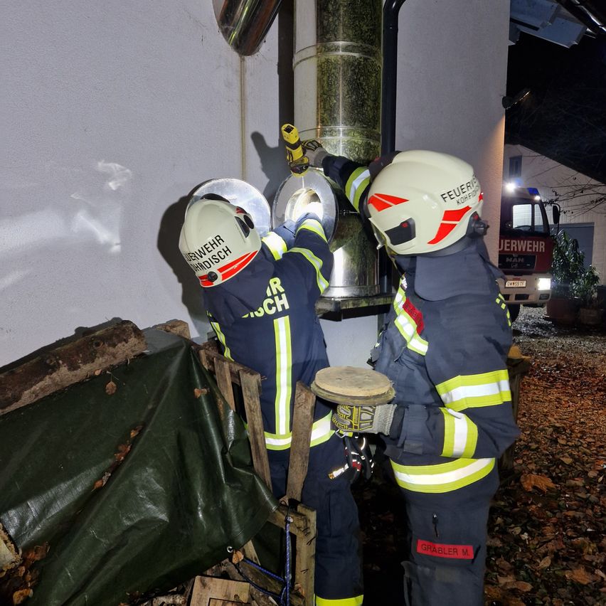 Zwei Feuerwehrleute arbeiten an einem Schornstein, einer benutzt ein Werkzeug am Schornstein, während der andere ein rundes Objekt in der Hand hält. Sie tragen beide Helme und Uniformen. Ein Feuerwehrwagen ist in der Nähe geparkt.